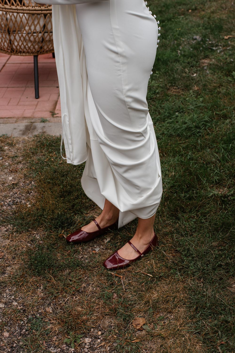 Lower half view of someone wearing flowing white pants and burgundy mary jane shoes standing on grass with a wicker chair visible.