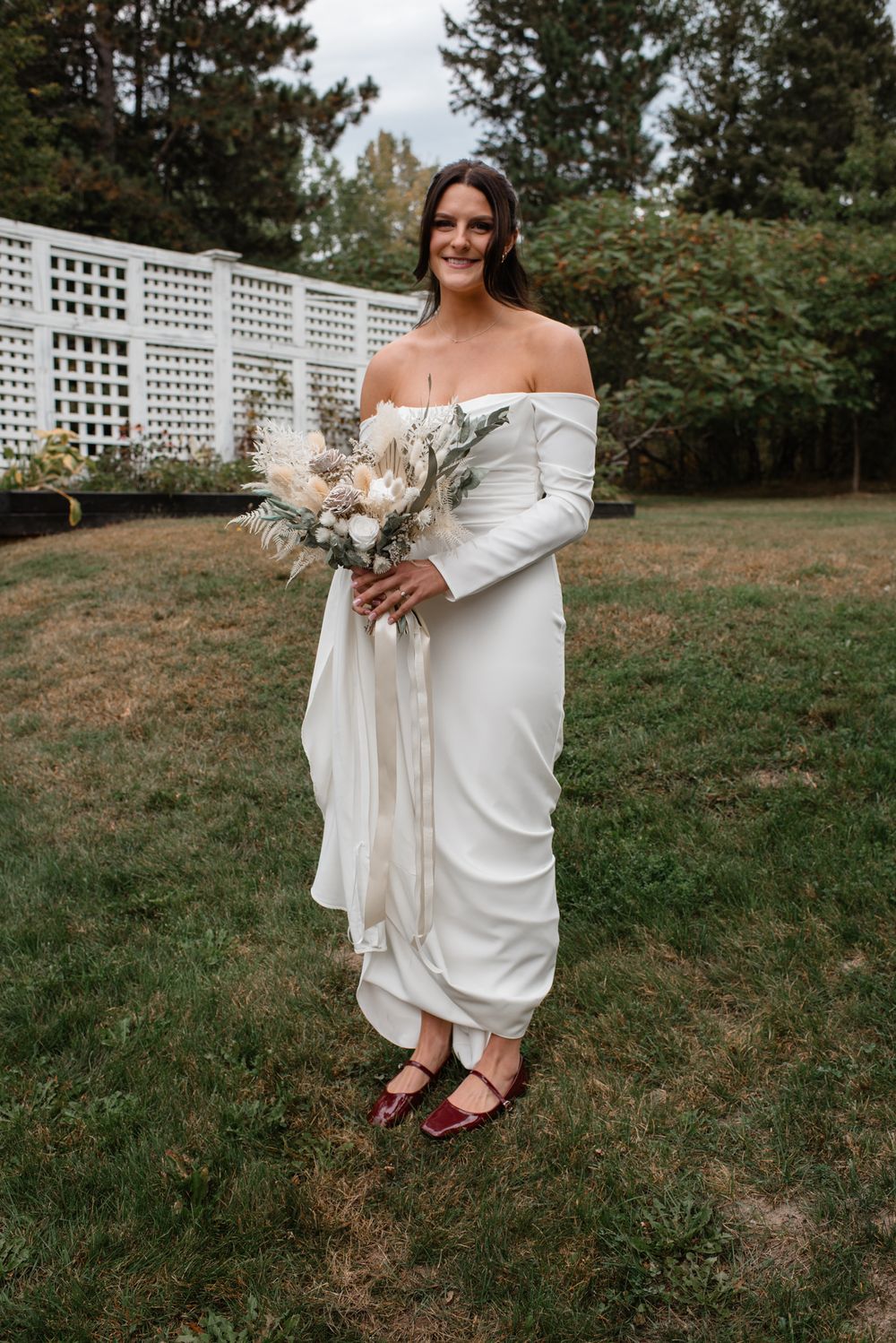 Elegant off-shoulder white wedding dress with long sleeves and neutral bouquet in outdoor garden setting.