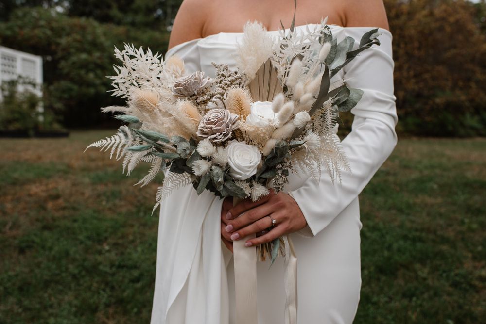 A rustic dried flower wedding bouquet featuring pampas grass and neutral tones held against a white dress outdoors.