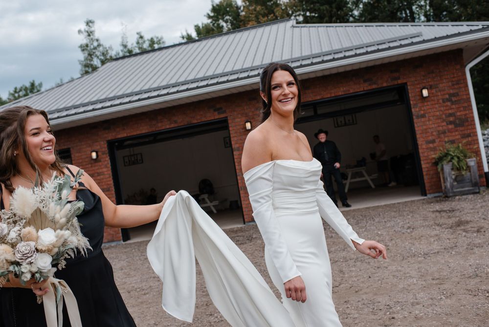Someone helps with flowing white wedding dress as they walk together outside a brick building with a gray metal roof.