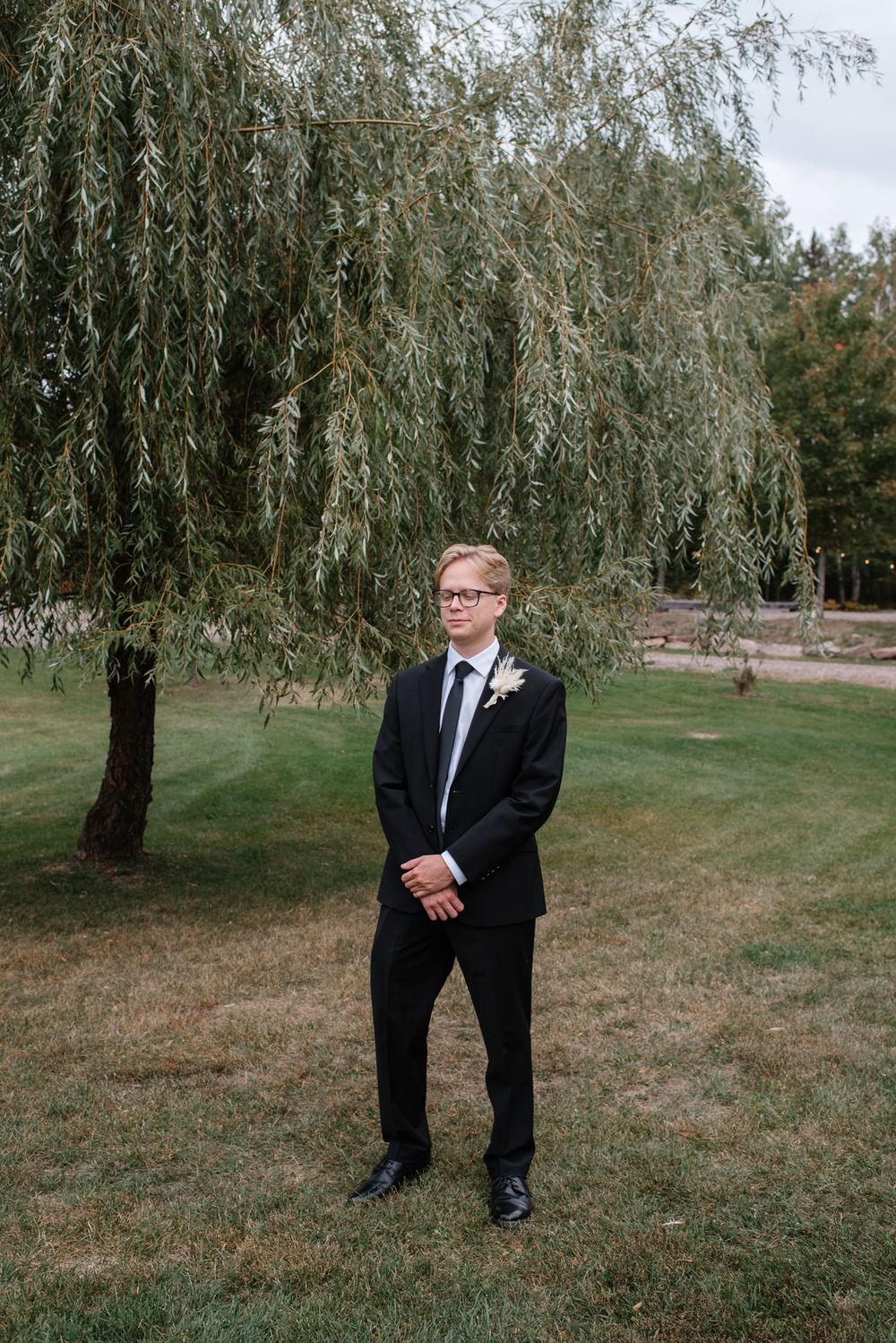 A figure in a formal black suit and tie stands beneath a graceful weeping willow tree on a manicured lawn.