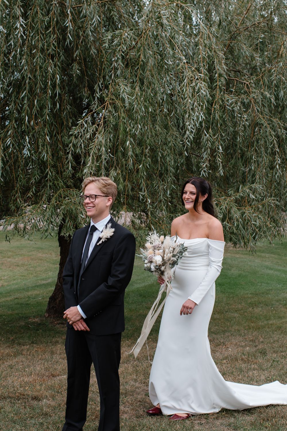 A couple in wedding attire standing together under a weeping willow tree on their special day.