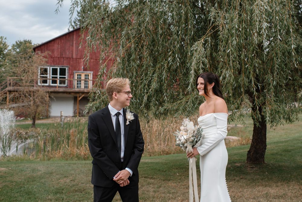 A couple shares a moment under a weeping willow tree in front of a rustic red barn during their wedding ceremony.