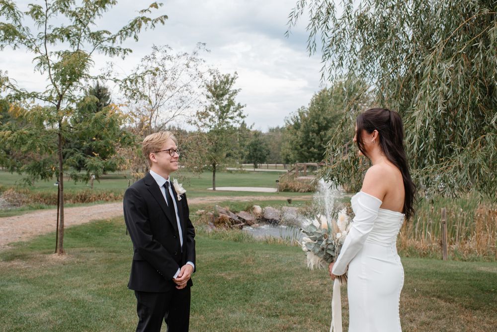A couple in formal wedding attire stands together outdoors near a pond with weeping willows and greenery.