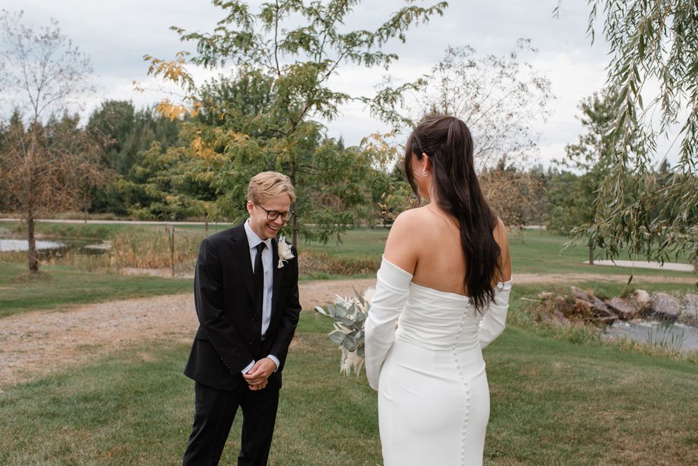 Two people in wedding attire share a candid moment together on a grassy lawn surrounded by autumn trees.