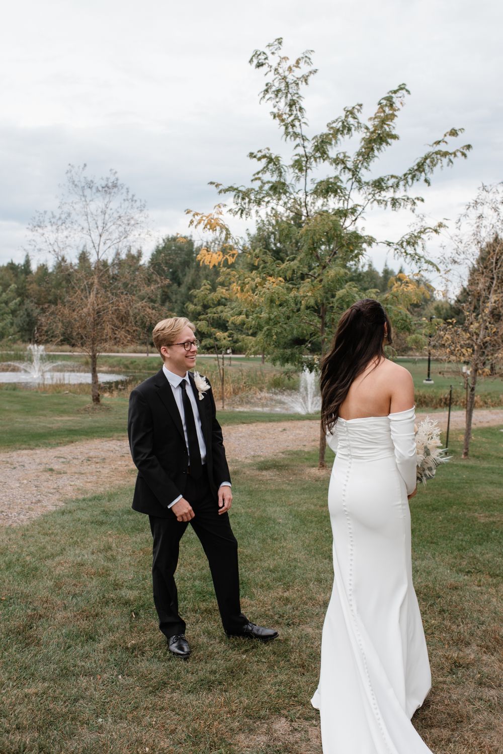 A couple in formal wedding attire stands on a grassy lawn with autumn trees and a lake in the background.