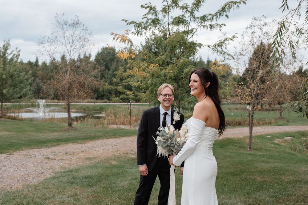 A couple stands together outdoors at a wedding ceremony on a gravel path surrounded by autumn trees and greenery.