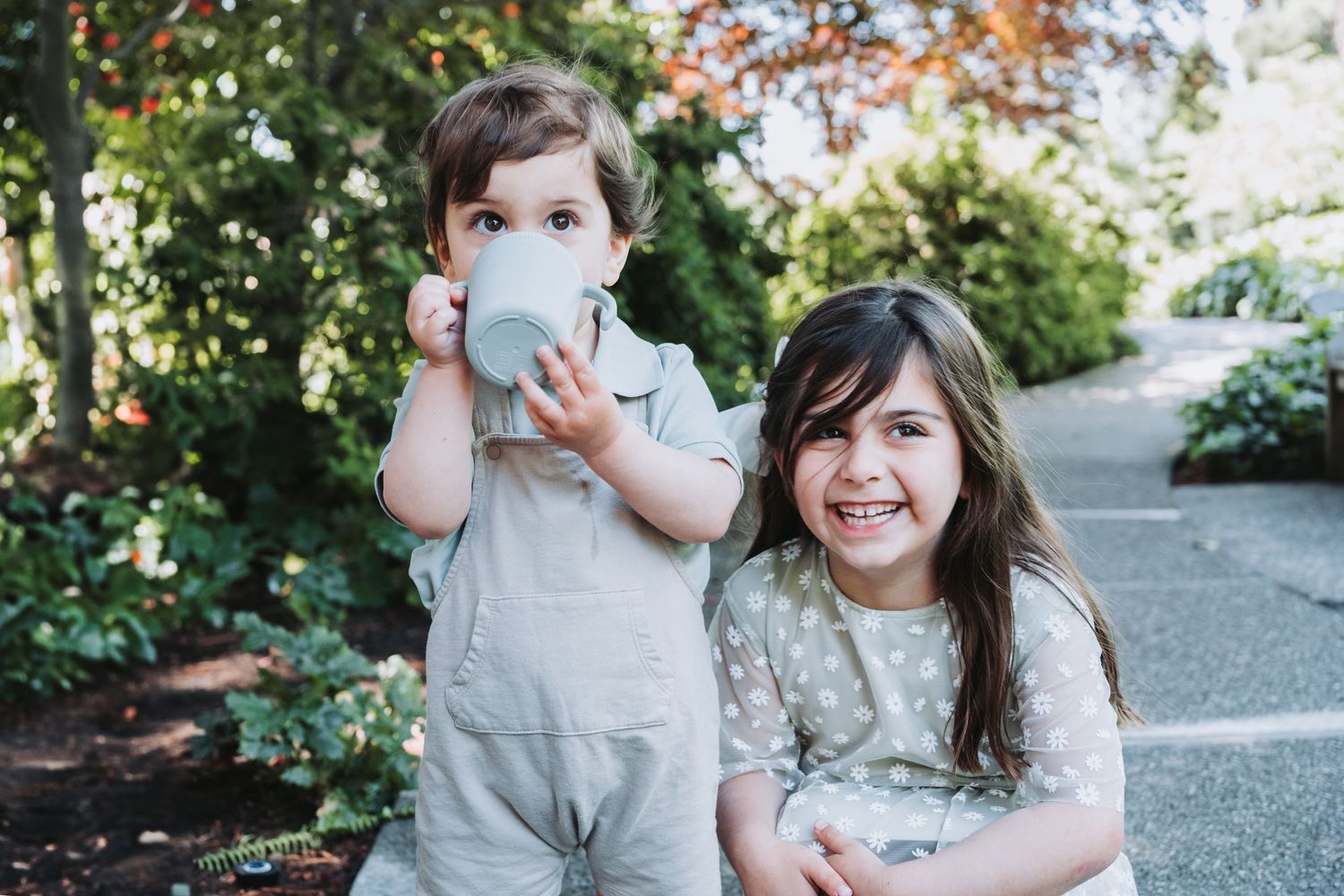 Parent and young child in grey overalls share a cheerful outdoor moment together while enjoying drinks in a garden setting.