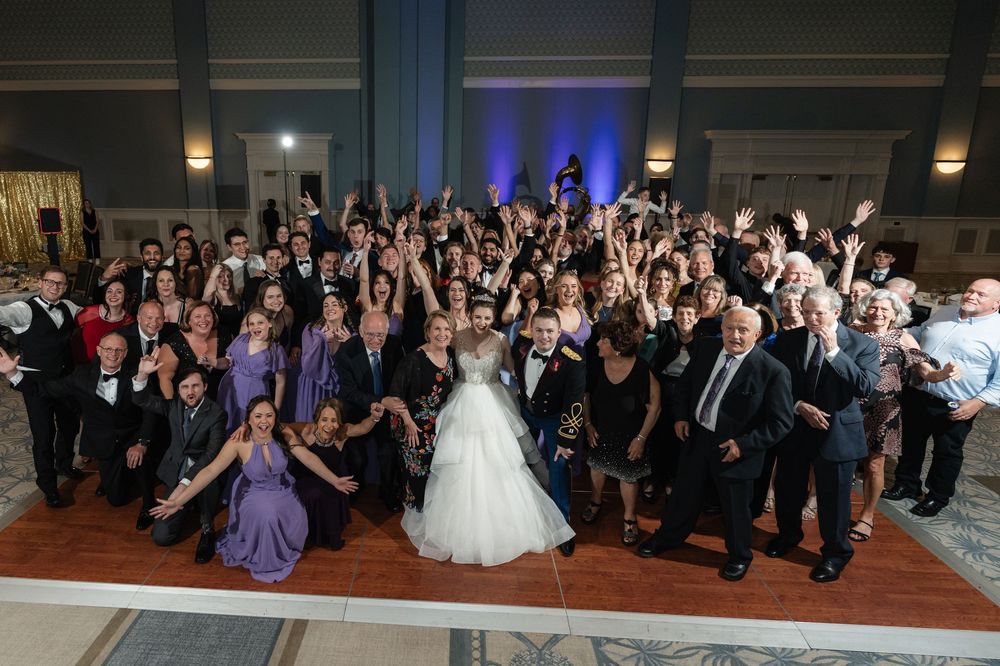 Large wedding reception group poses together on dance floor with arms raised in celebration at elegant venue with blue lighting.