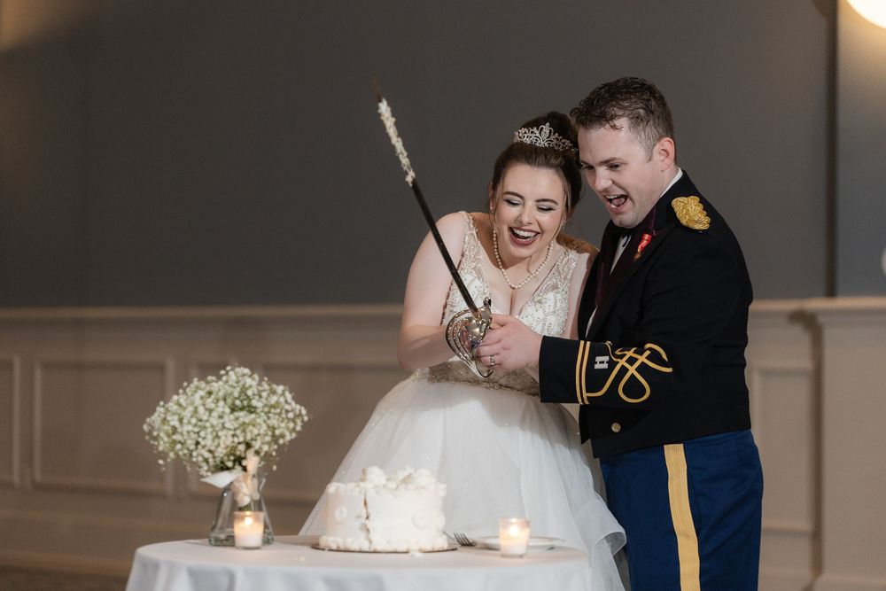 A joyful couple cuts their wedding cake together while wearing military dress uniform and white wedding gown.