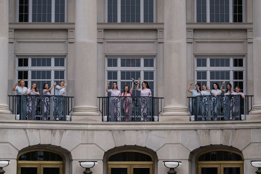 Groups of people stand on three ornate stone balconies of a classical building with arched doorways below.