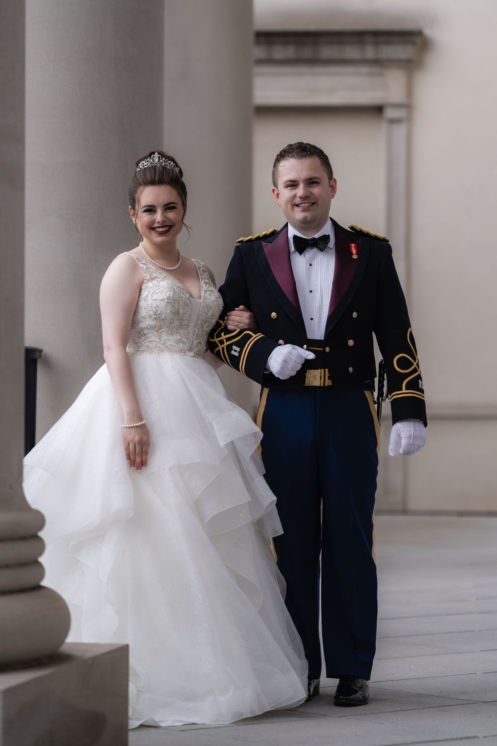 A wedding couple poses in formal attire, with the bride in a layered white gown and the groom in military dress uniform.