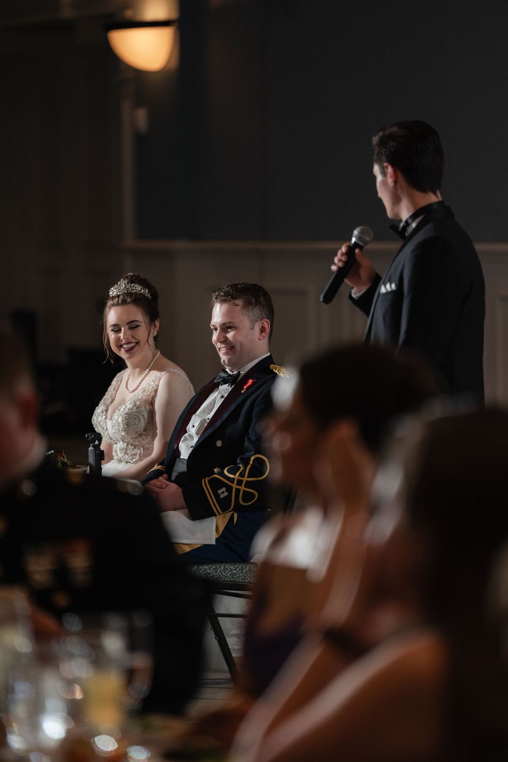 Military wedding reception scene in dimly lit venue with happy couple listening to speaker during toasts.
