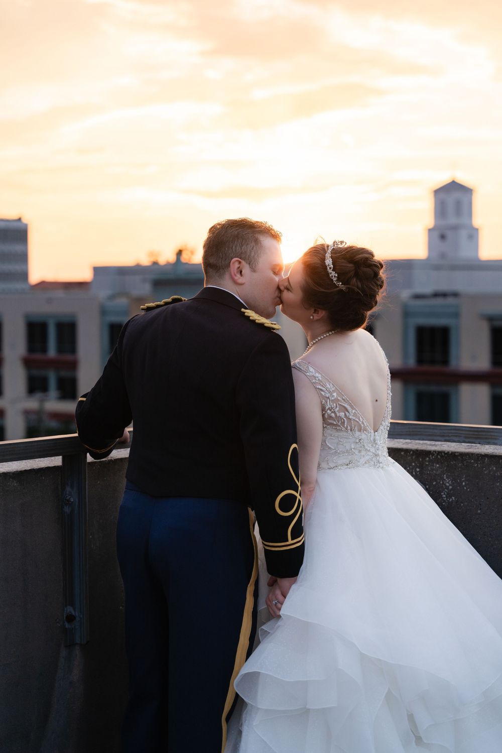 A romantic sunset kiss on a rooftop with a person in formal military attire and someone wearing a white layered wedding gown.