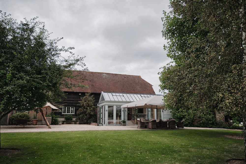 A rustic barn wedding venue with a modern glass conservatory addition and manicured lawn on a cloudy day.