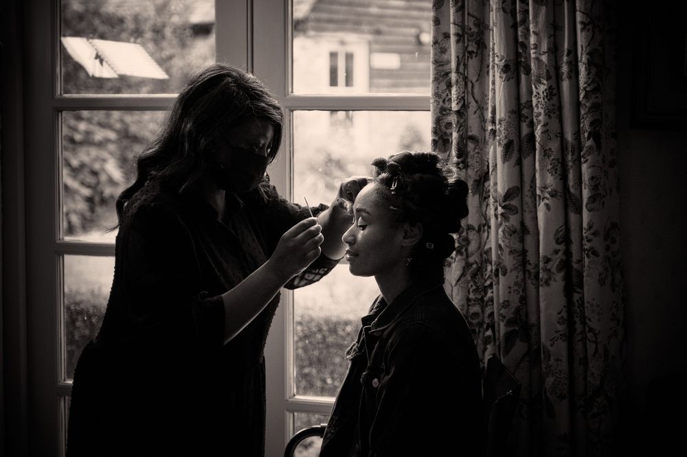 A silhouette of someone applying makeup near a window with natural light streaming in, creating a moody black and white scene.