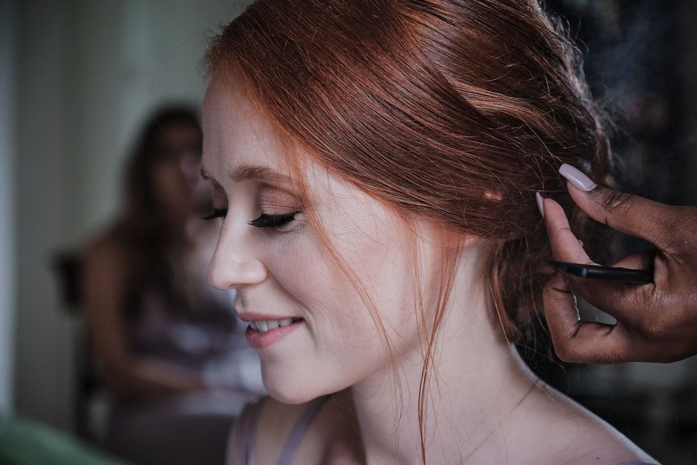 A woman smiles gently while having her hair styled, captured in soft natural lighting during wedding preparations.