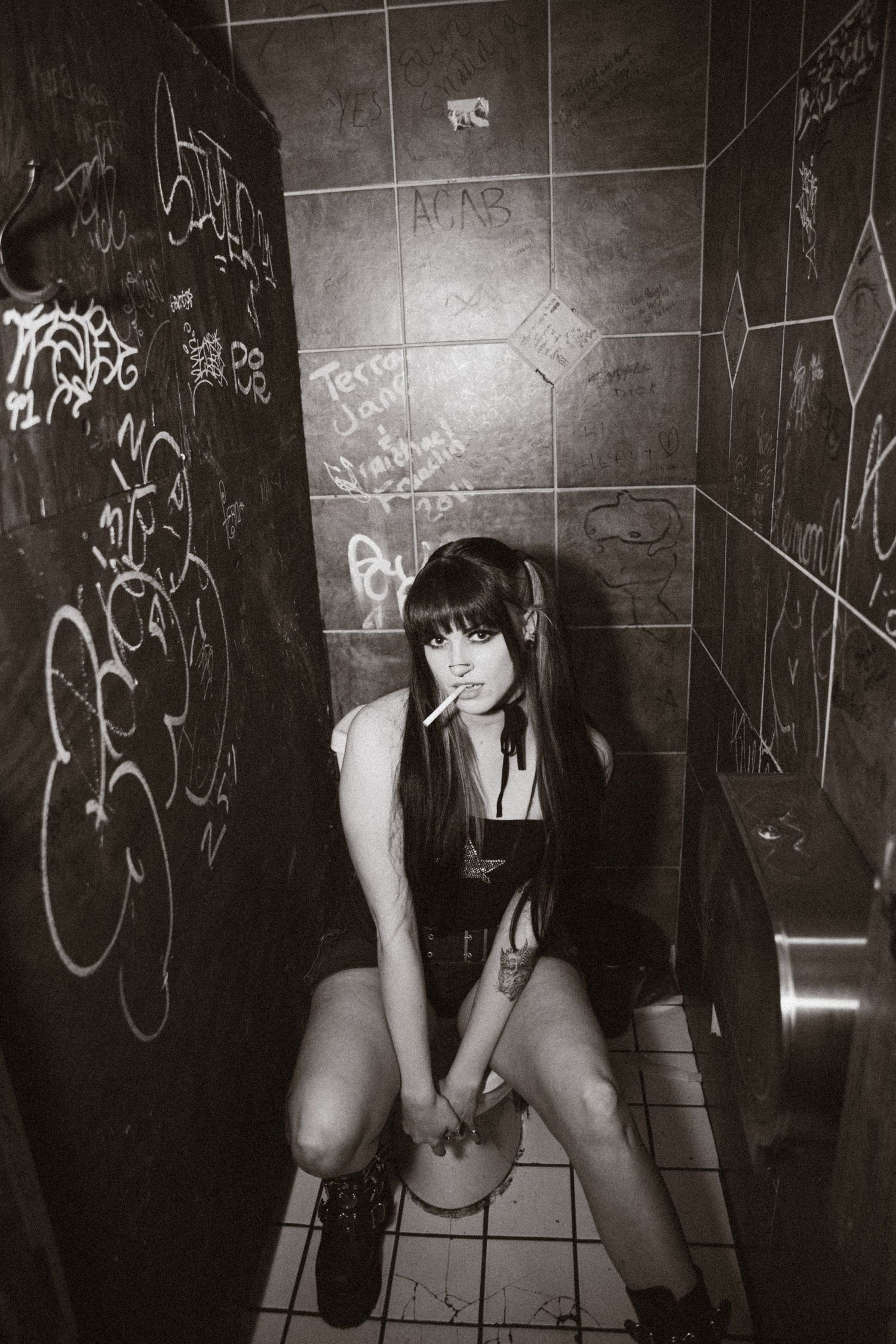 A black and white photograph taken in a graffiti-covered bathroom stall with tiled walls and floor.