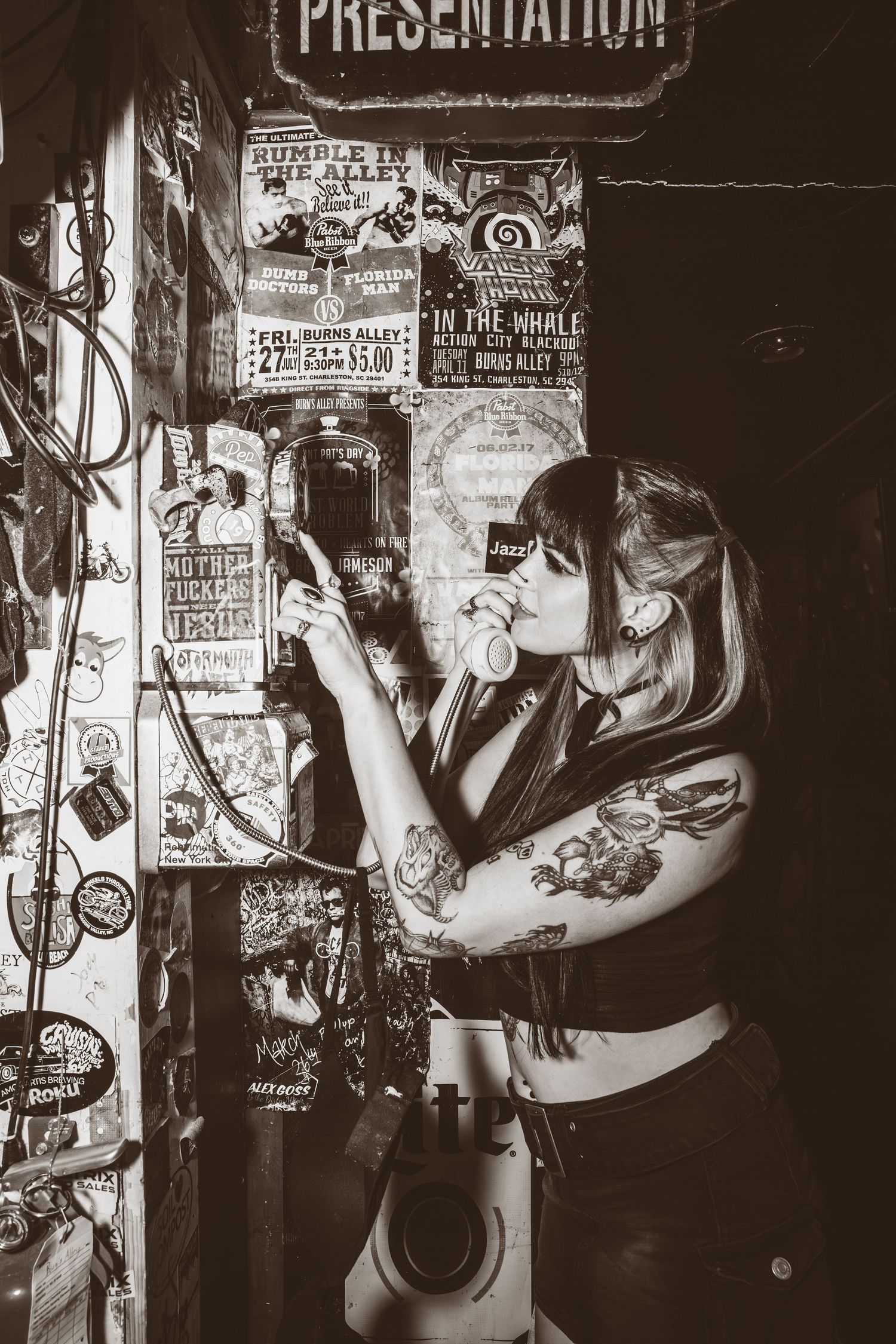 Black and white photo of a tattooed person in punk rock style posting flyers on a wall covered in show posters.