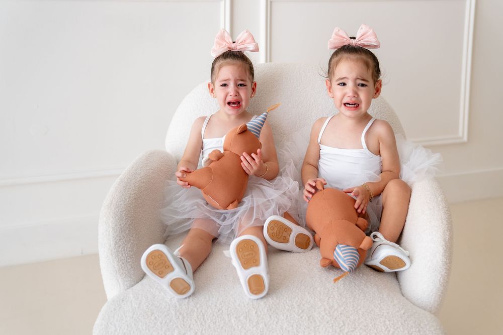 Two toddlers in white tops and gray bottoms sit on white furniture while holding orange pumpkins.