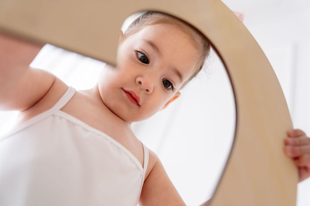 Dancer in white leotard peeking through a curved wooden prop in bright studio setting.