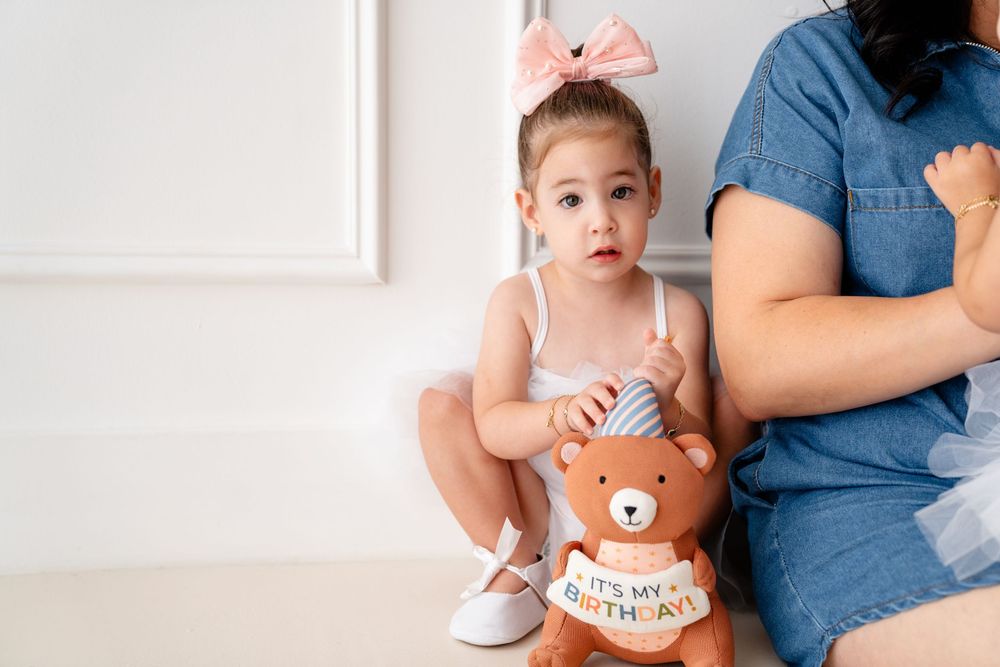 A toddler wearing a cute brown bear onesie sits next to someone in denim on a white floor.