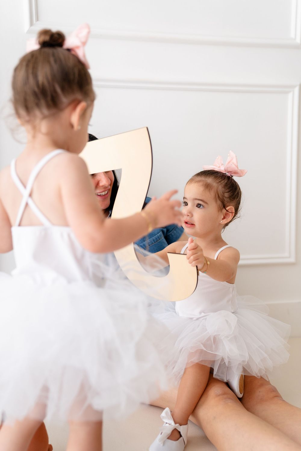 Young ballerina in white tutu holding a wooden number prop while practicing in a dance studio.