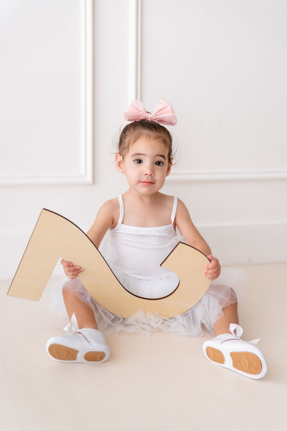 Young dancer in white tutu and ballet shoes holds a large decorative letter while sitting on white floor.