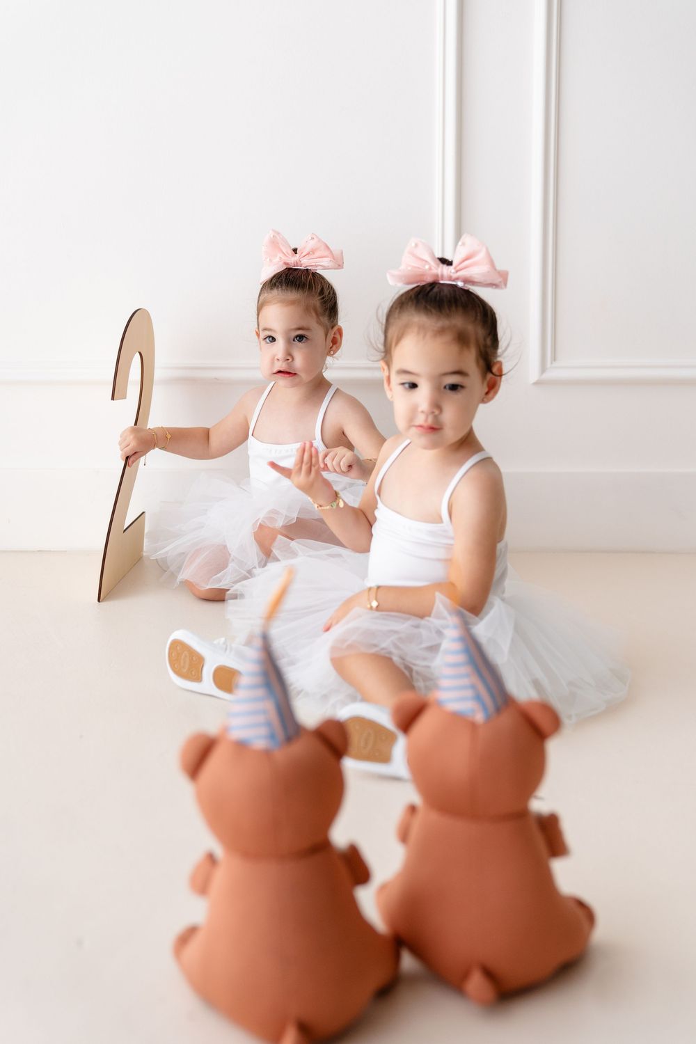 Two small dancers in white tutus and pink bows sit on a white floor with decorative ceramic bears.