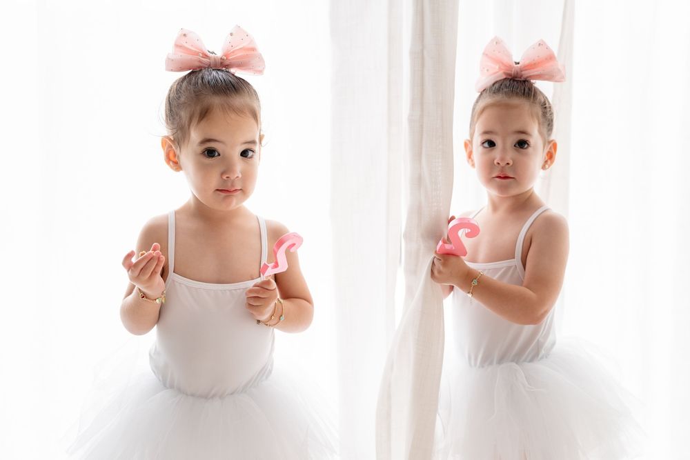 Two young ballet dancers in white tutus posing against light curtains with pink hair bows.
