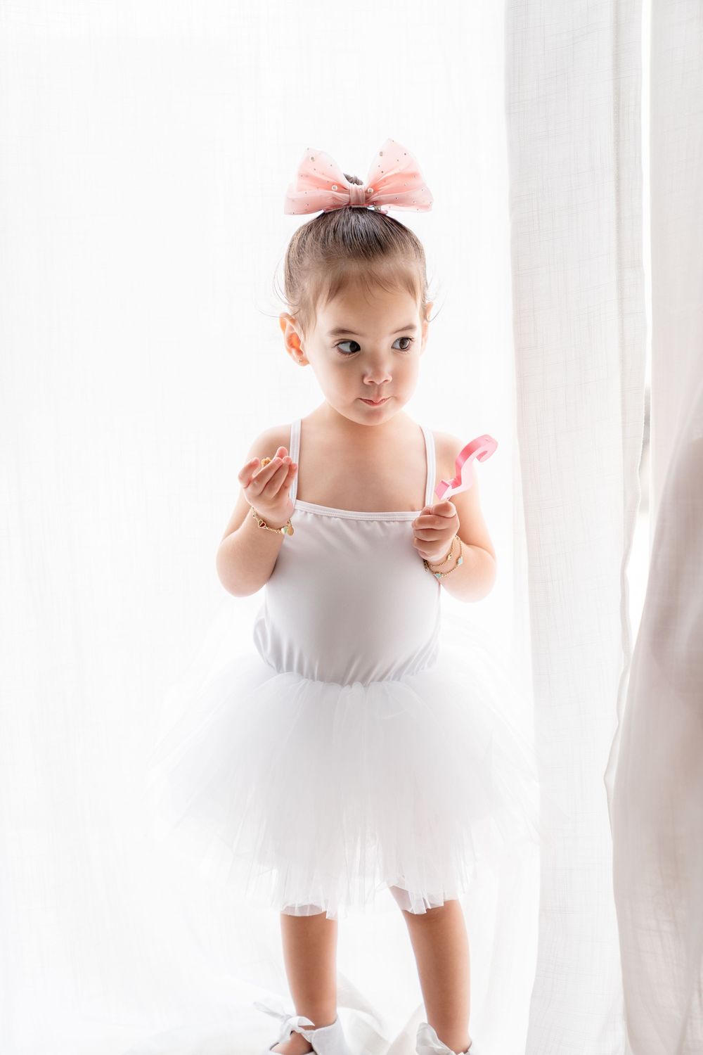 Young dancer in white tutu and pink bow poses gracefully near white curtains.
