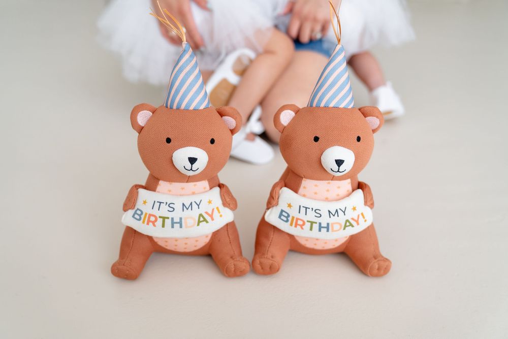 Two brown teddy bear cookies decorated with 'It's My Birthday' text sit next to baby feet wearing white shoes.