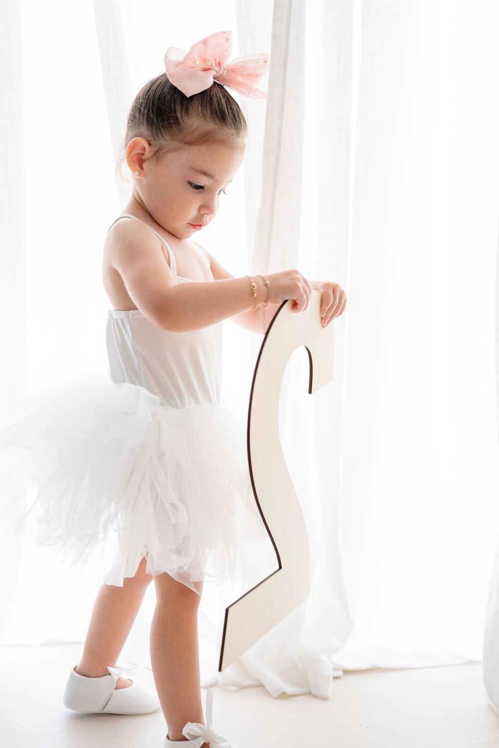 Young ballet dancer in white tutu and pink bow practicing poses near a bright window.