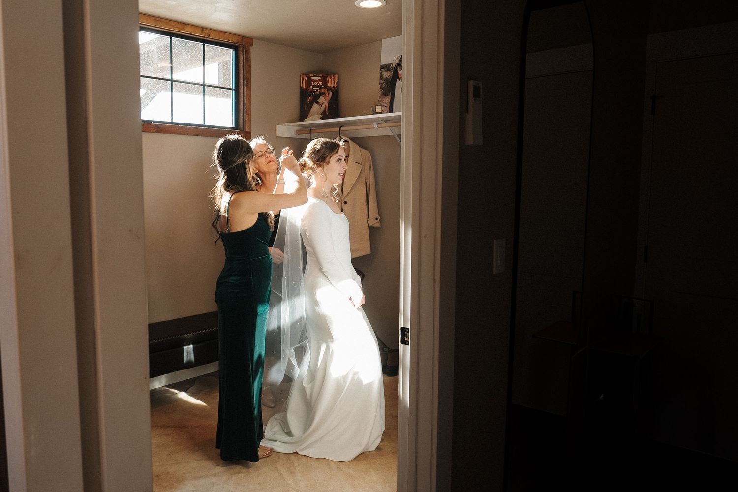 Two people prepare formal attire in a small dressing room with warm lighting and a shelf on the wall.
