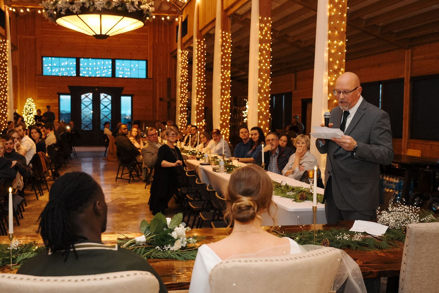 A speaker addresses guests during an elegant indoor wedding reception with string lights and wooden beams overhead.