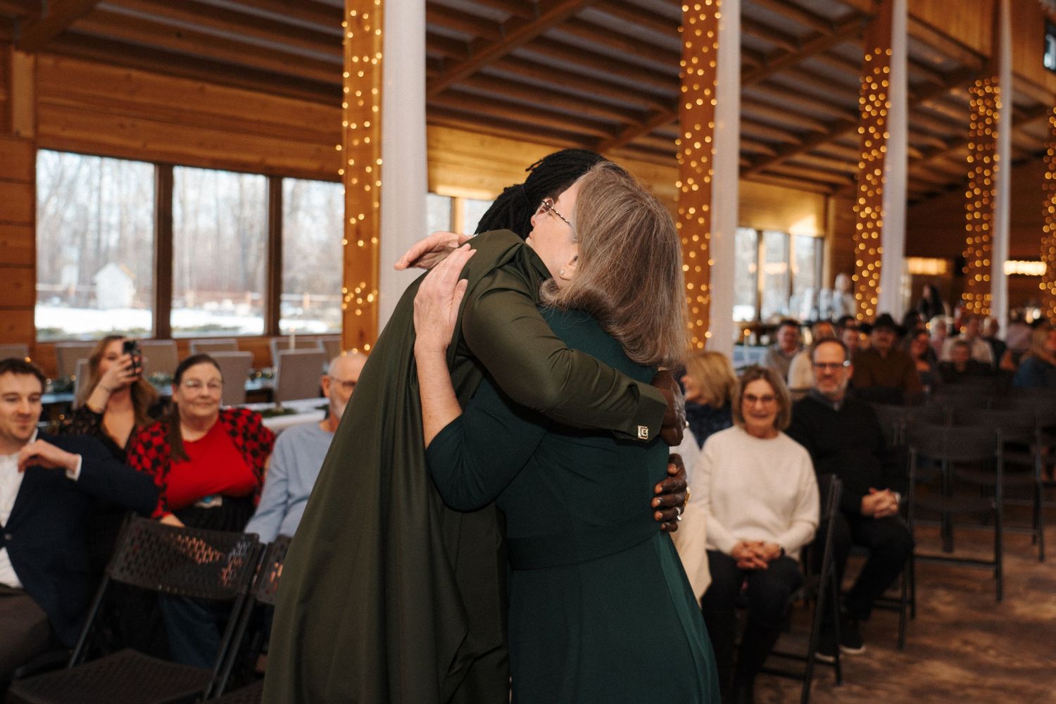 Two people in green dresses embrace during a winter ceremony with string lights and guests watching in wood-paneled room.