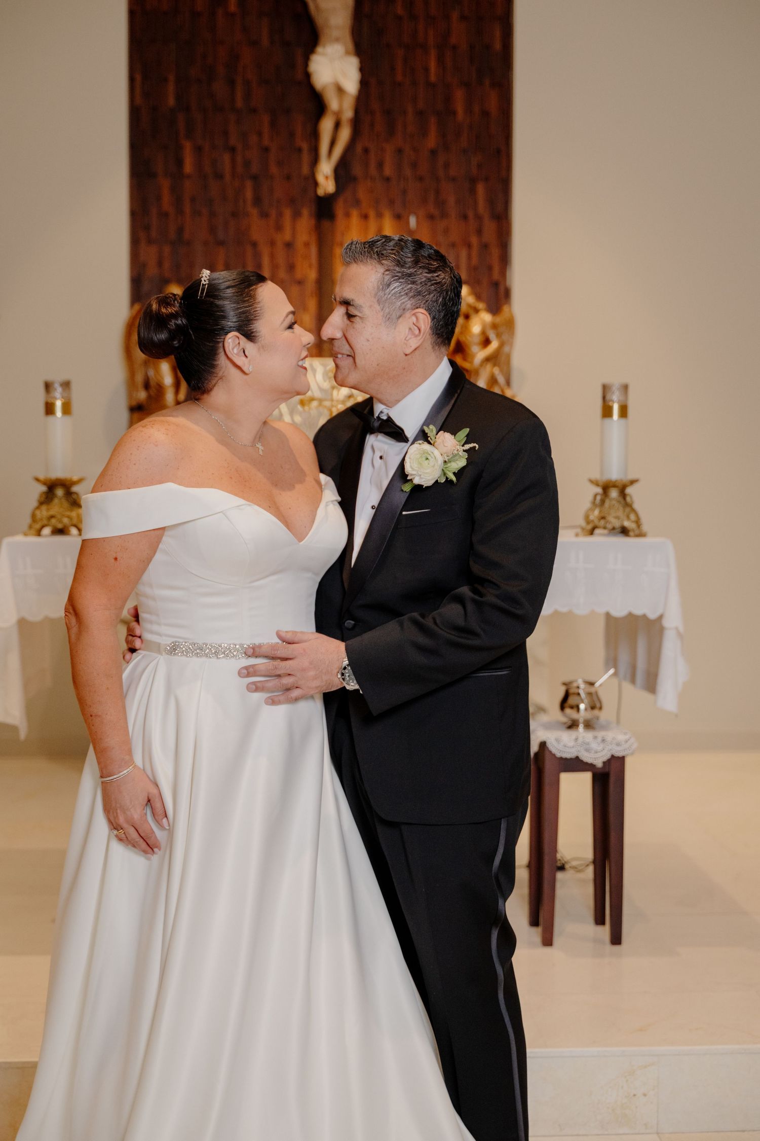 A loving couple in wedding attire share an intimate moment in front of a church altar with candles and crucifix.