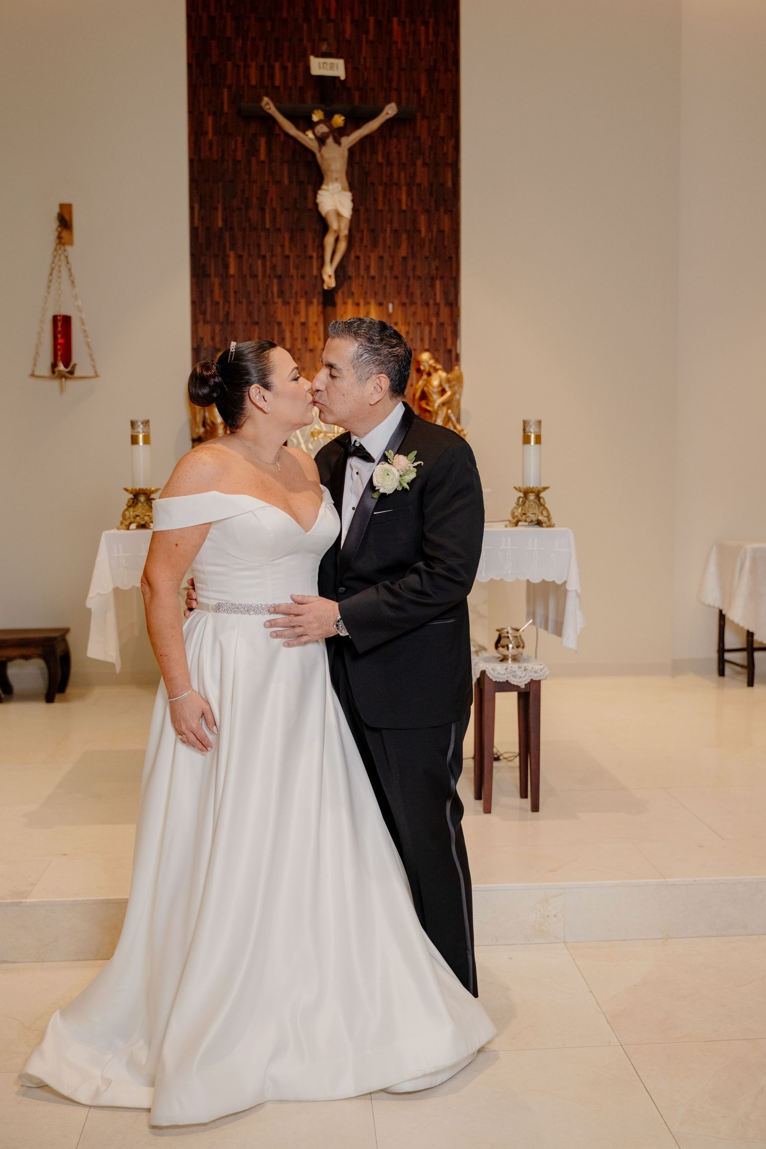 Newlyweds share an intimate moment in a church with a crucifix mounted on the textured wall behind them.