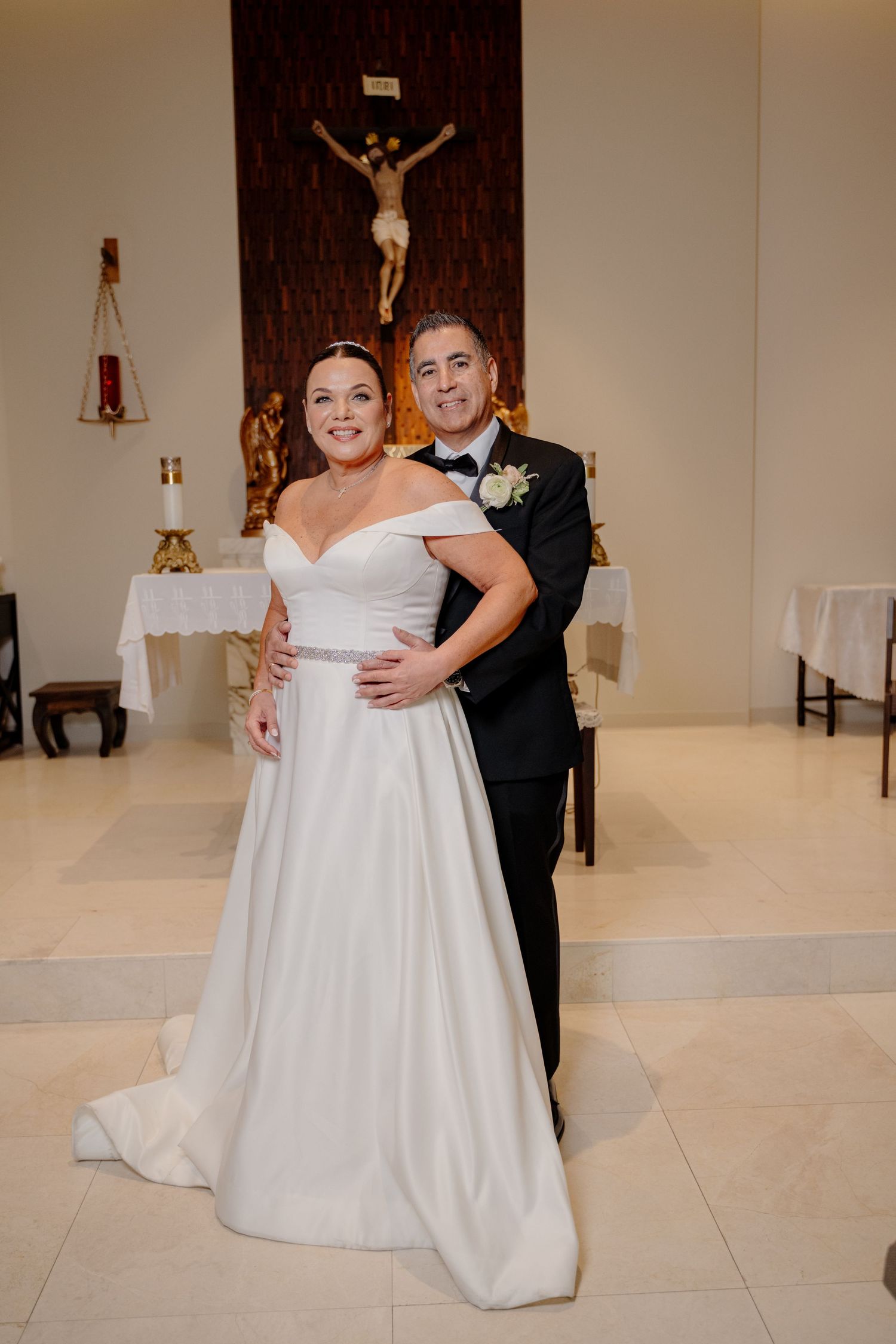 A wedding couple poses together in a church with a crucifix on the wall behind them.