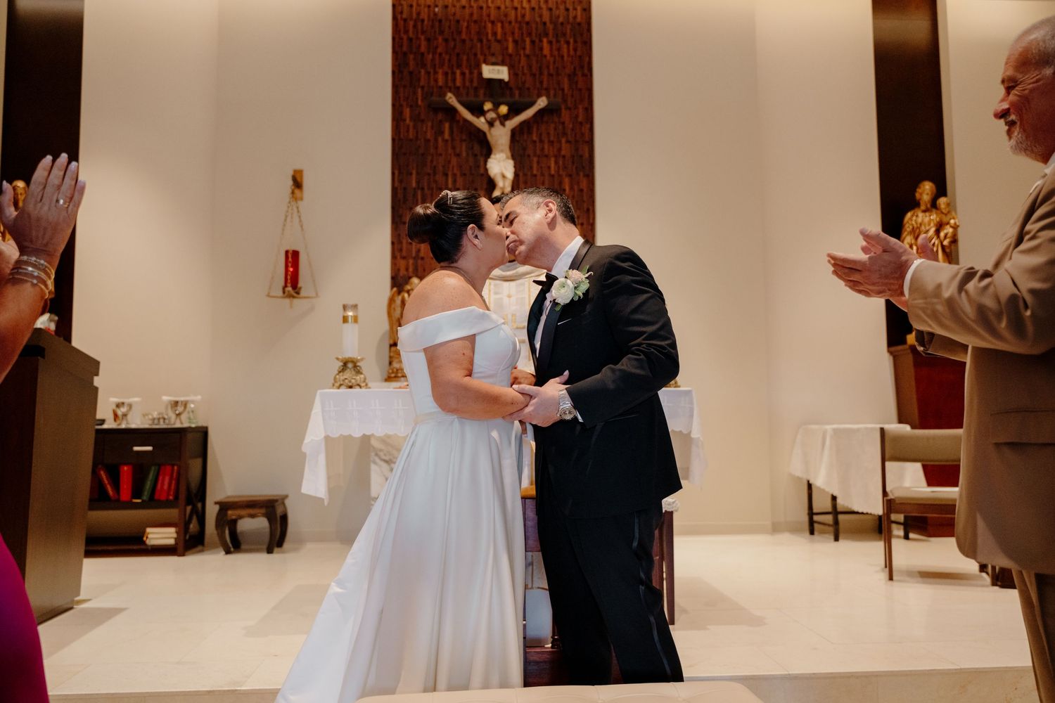 A bride and groom share a tender moment at the altar in a Catholic church with a large crucifix mounted on the wall.