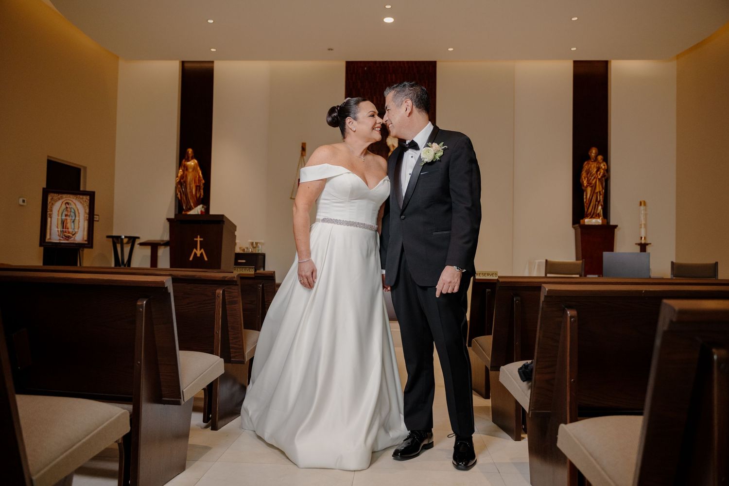 A couple shares an intimate moment together in their wedding attire inside a chapel with wooden pews.