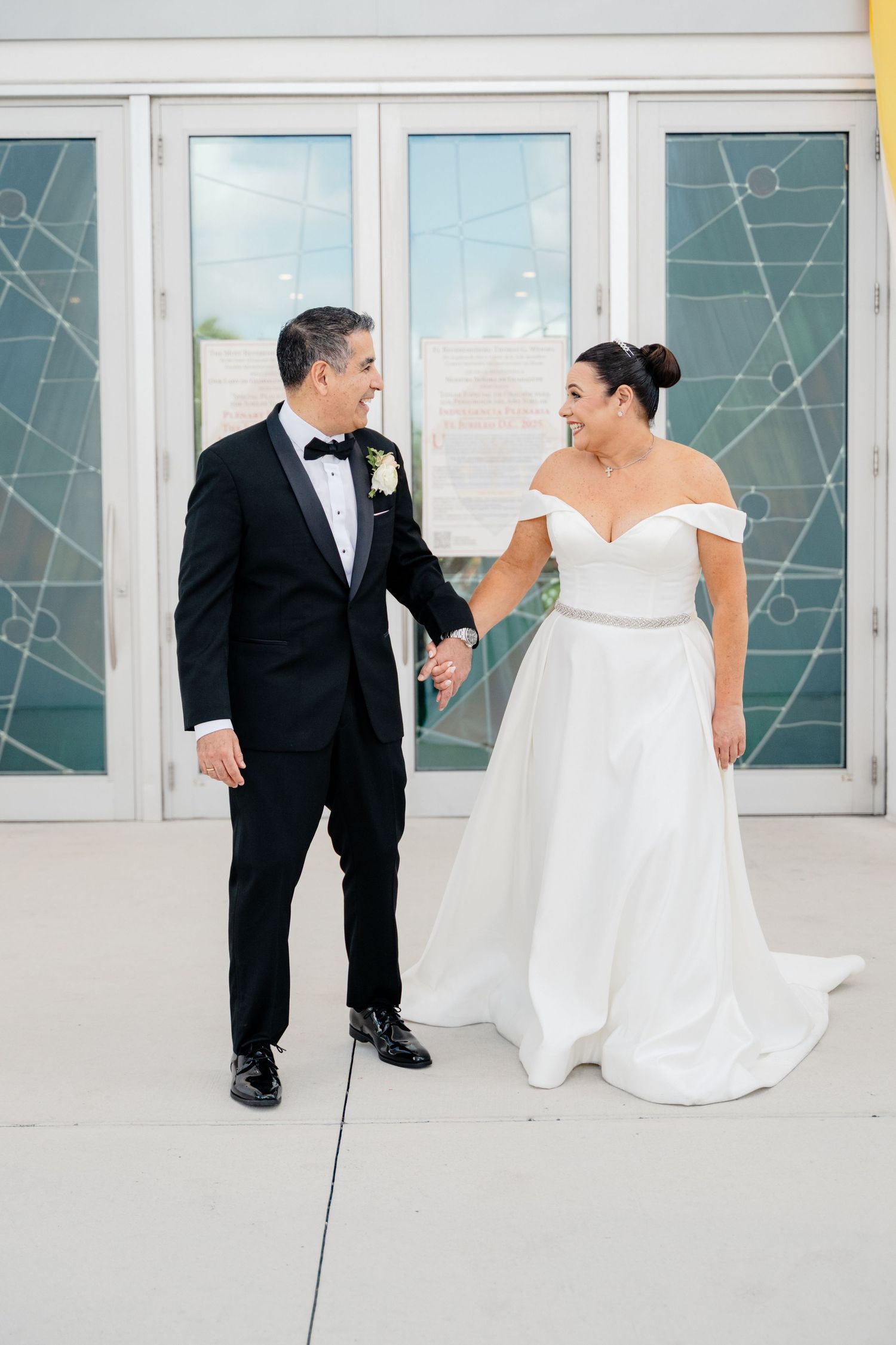 Bride in off-shoulder white gown and groom in black tuxedo hold hands while laughing together against glass wall exterior.