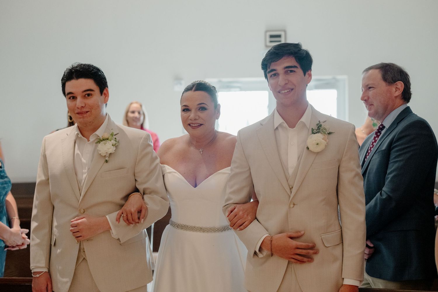 Three wedding attendees walk arm in arm, with two in tan suits flanking a person in a white off-shoulder dress.