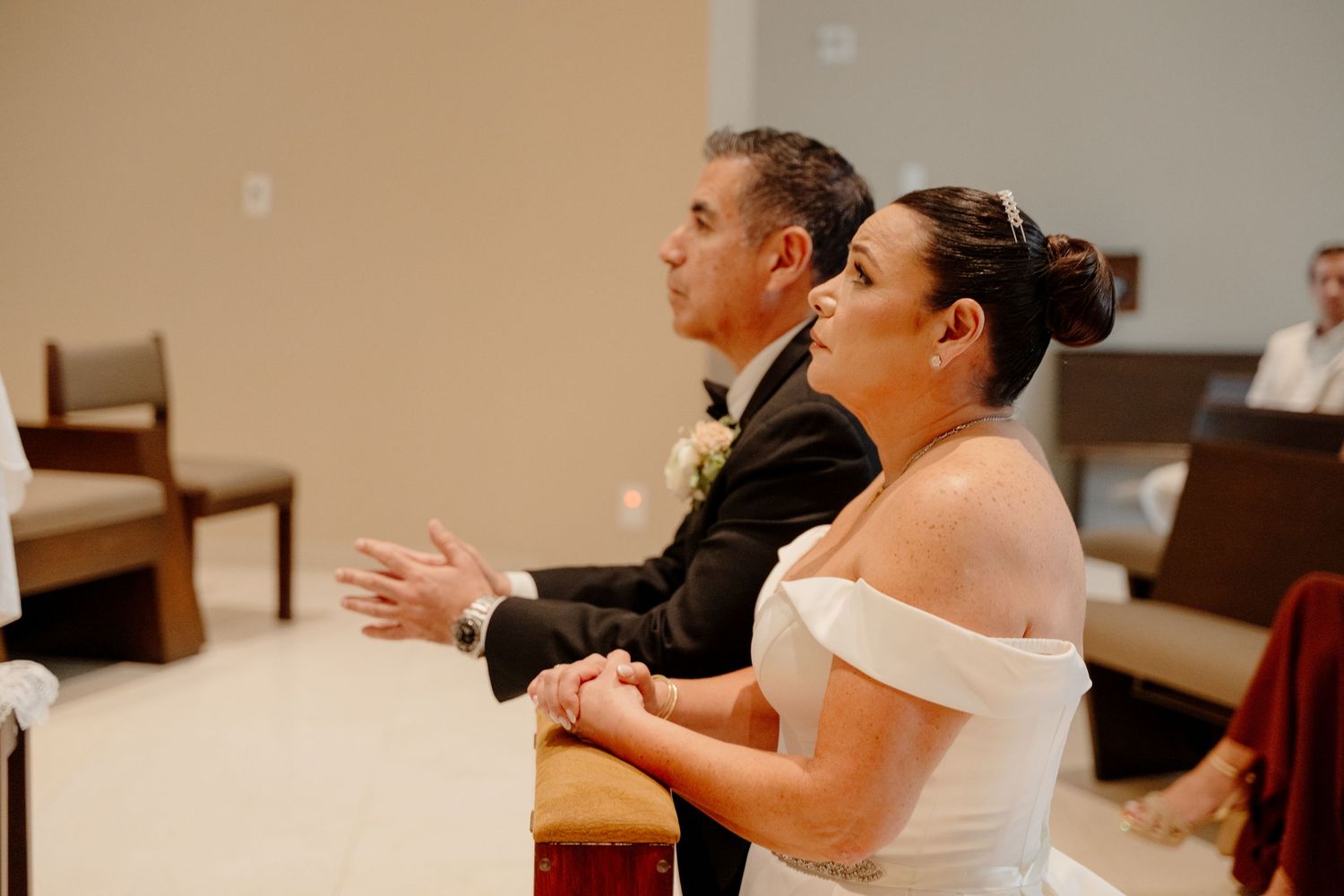 A couple in formal wedding attire sits together in a church setting with their hands clasped in prayer.