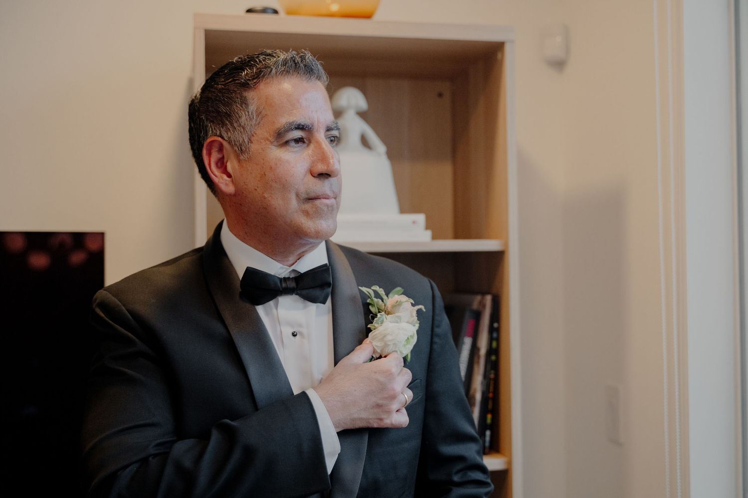 A person in a black tuxedo with bowtie and white boutonniere adjusts their lapel while getting ready.