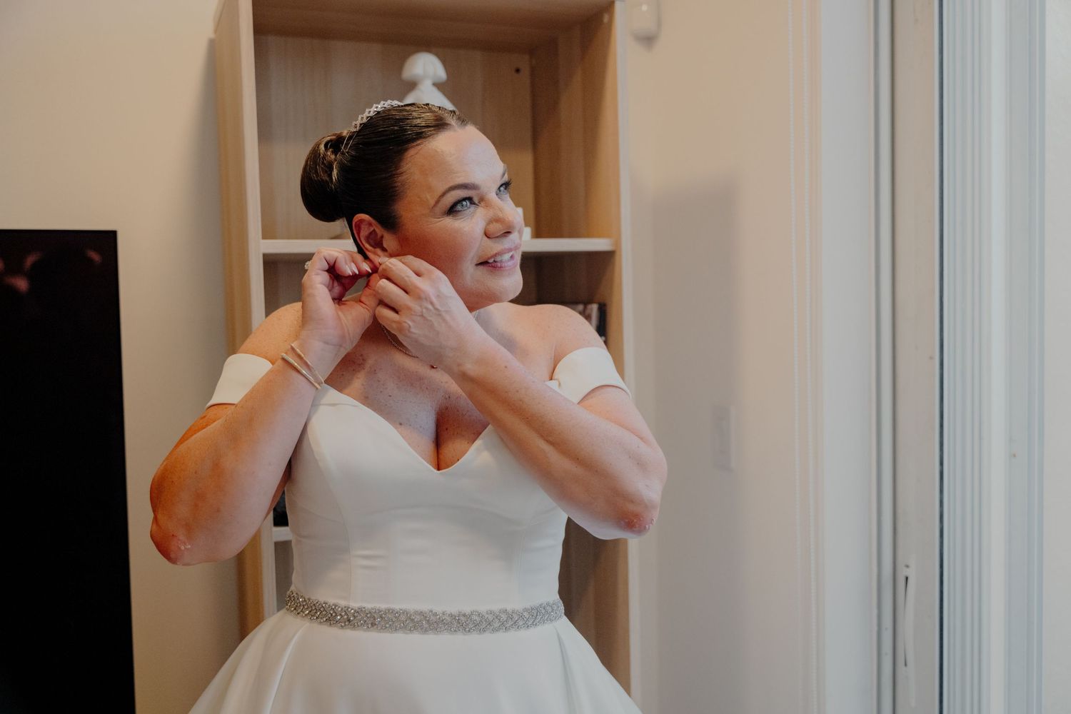 A bride in a classic white wedding gown with a beaded belt puts on earrings while getting ready on her wedding day.