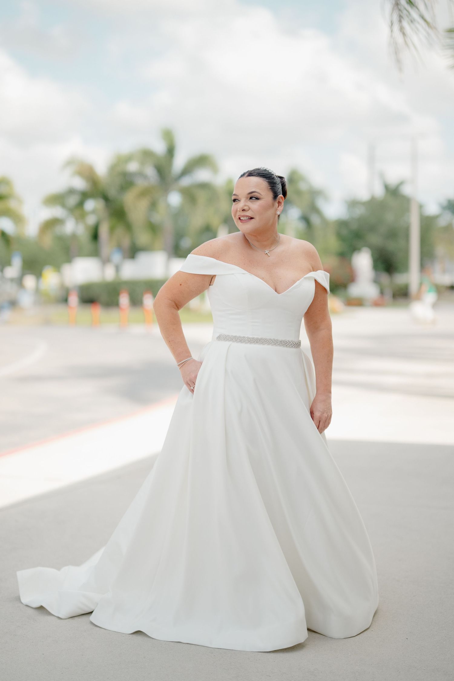 An elegant off-shoulder white wedding gown with a flowing A-line silhouette and silver belt detail against a tropical backdrop.