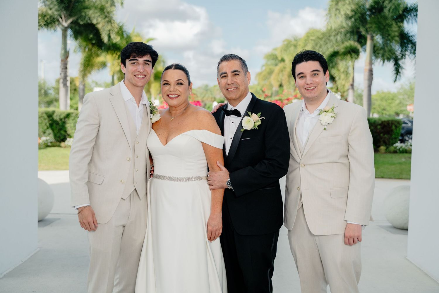 A wedding party poses together on white walkway with palm trees in background at outdoor venue.