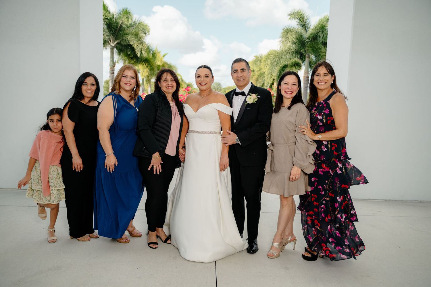 A group of people in formal attire posing together at a wedding celebration with palm trees in the background.
