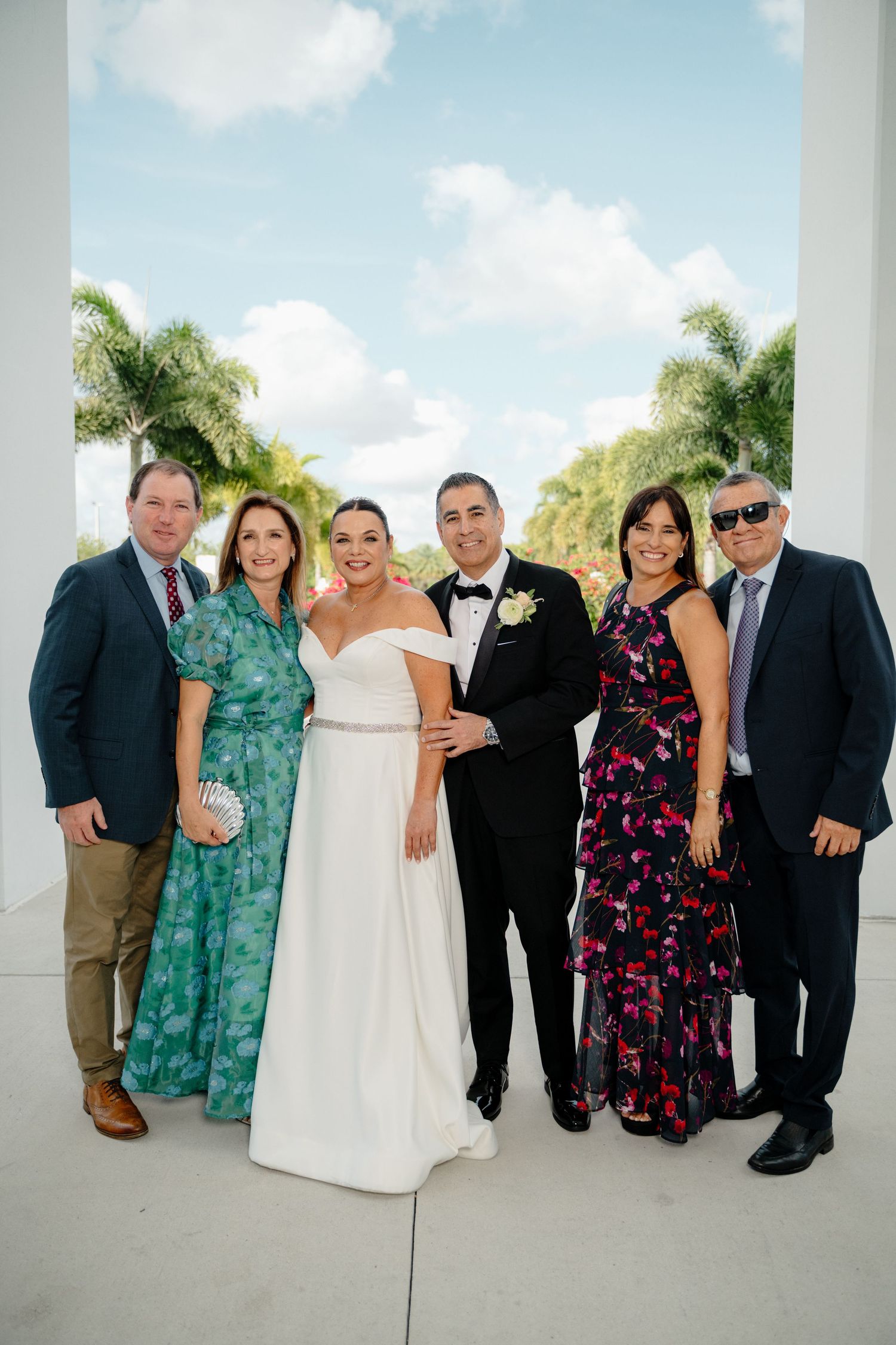 Wedding party poses together on a sunny day with palm trees in background at an elegant white venue.